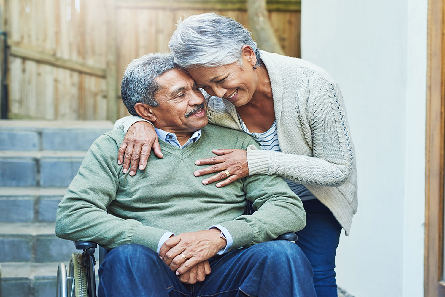 Older Latin couple with gray/salt pepper hair color hugging. Man is sitting down in a wheelchair with a green sweater on. Women is standing above him in a cream colored sweater